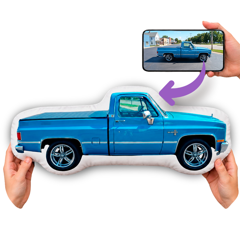 A custom printed throw pillow shaped like a bright blue pickup truck is held by hands against a white background. An inset phone screen at the top shows the source photo used for the custom print, with a purple arrow pointing to the pillow.